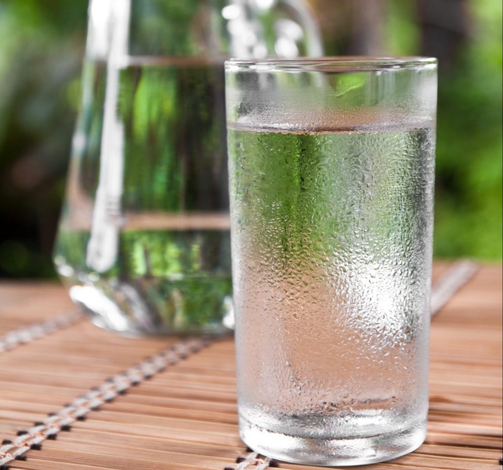 Kaltes Glas Wasser mit einer Kanne Wasser im Hintergrund. Auf einem Tisch im Freien.