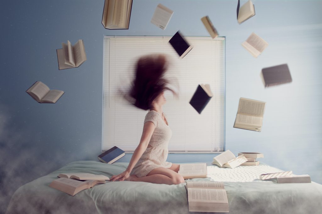 Frau sitzt auf Bett mit etlichen Büchern, welche um sie herum fliegen