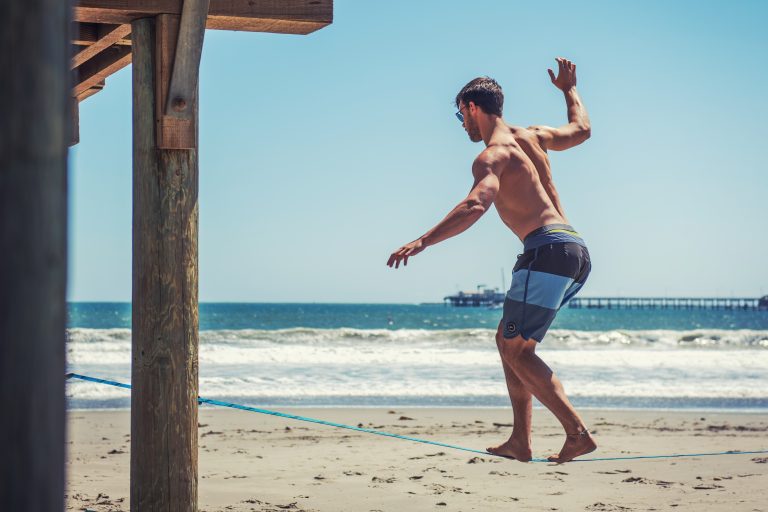 Mann am Strand balanciert auf der Slackline. Im Hintergrund ist das Meer zu sehen.